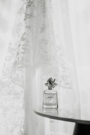 Black and white photo of Marc Jacobs Daisy perfume bottle on a glass table with lace curtains backdrop.