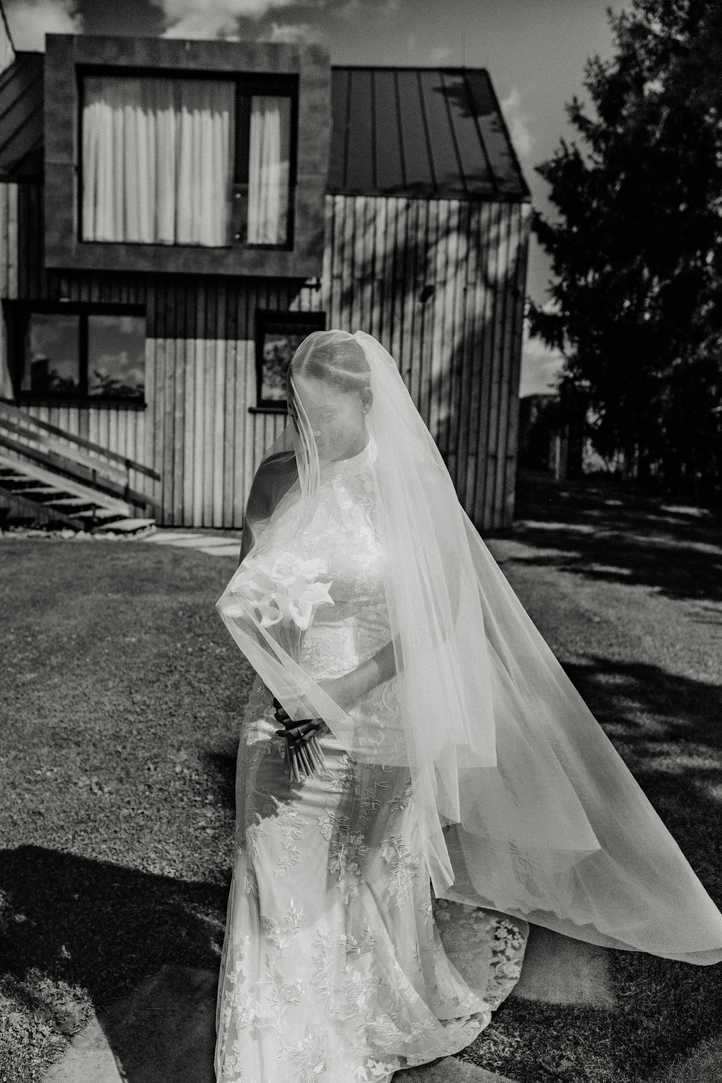 Black and white photo of a bride in lace gown with flowing veil, standing outside a modern building on her wedding day.