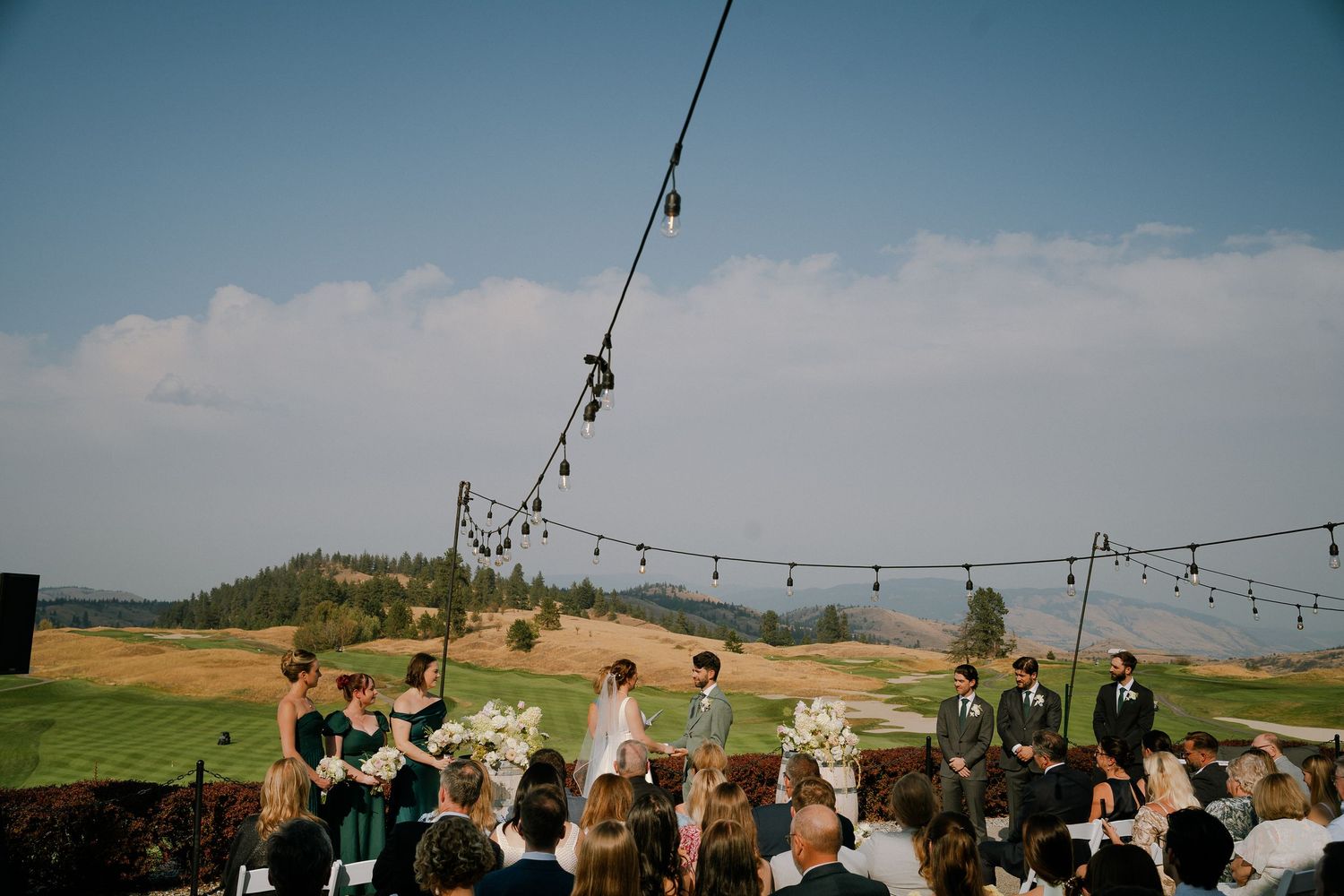 Outdoor wedding ceremony on a golf course with string lights, guests seated, and scenic green hills under a blue sky.