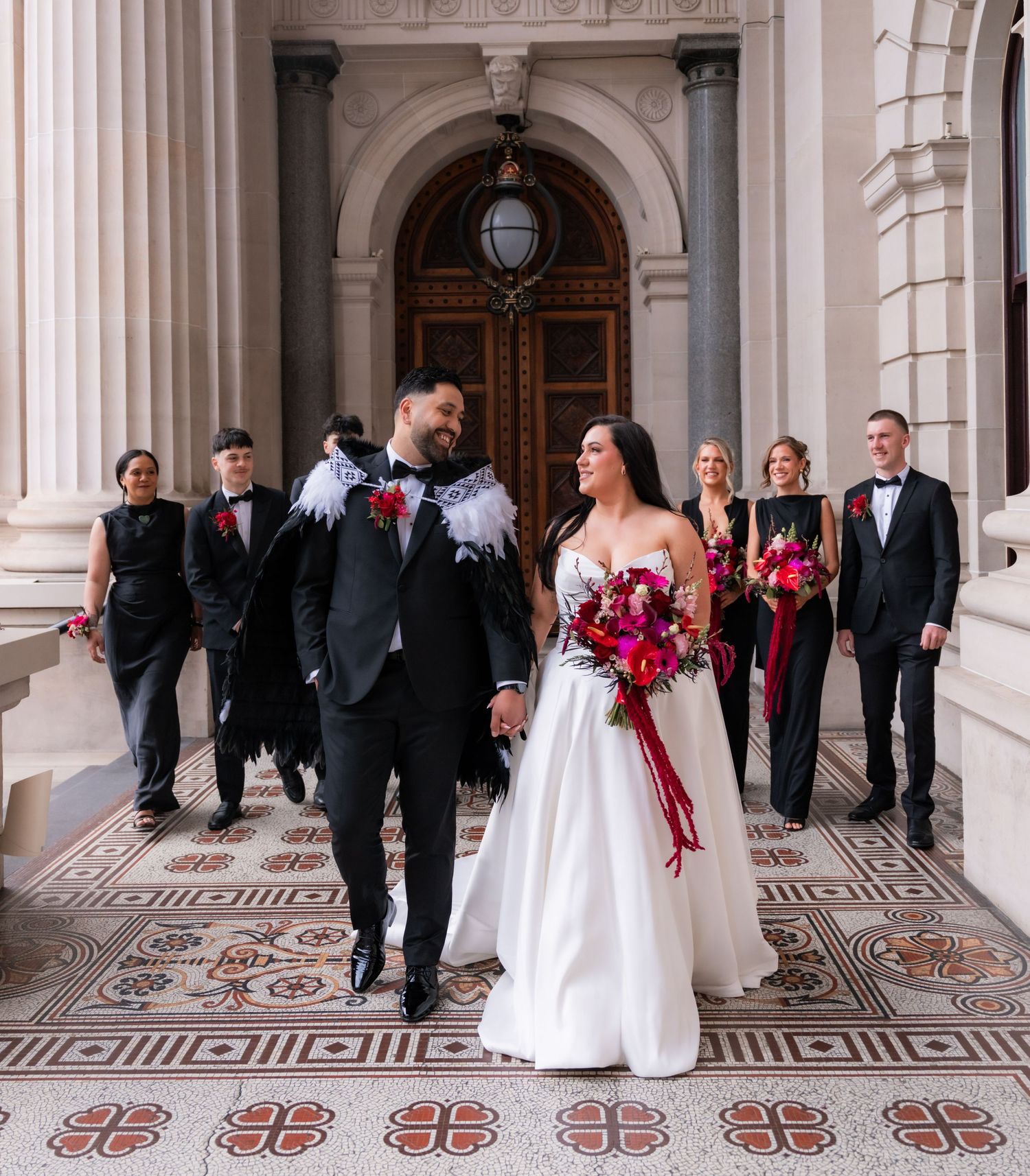 Bride and groom walk hand-in-hand at elegant venue with wedding party in black and burgundy attire behind them.
