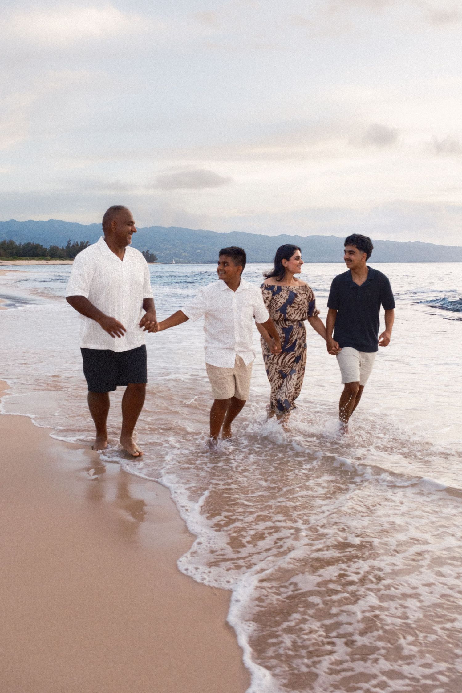 Family of four walking along a sandy beach shoreline with ocean waves and mountain backdrop at dusk.