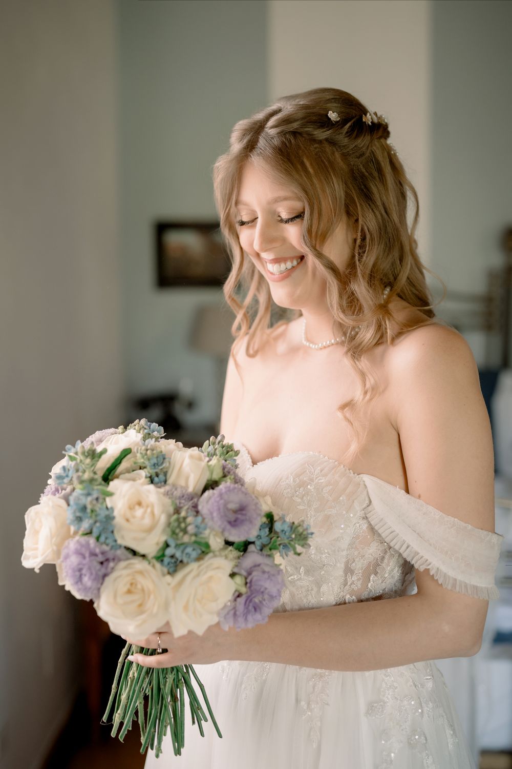 Smiling bride in off-shoulder gown holding a bouquet of white roses and purple flowers on her wedding day.