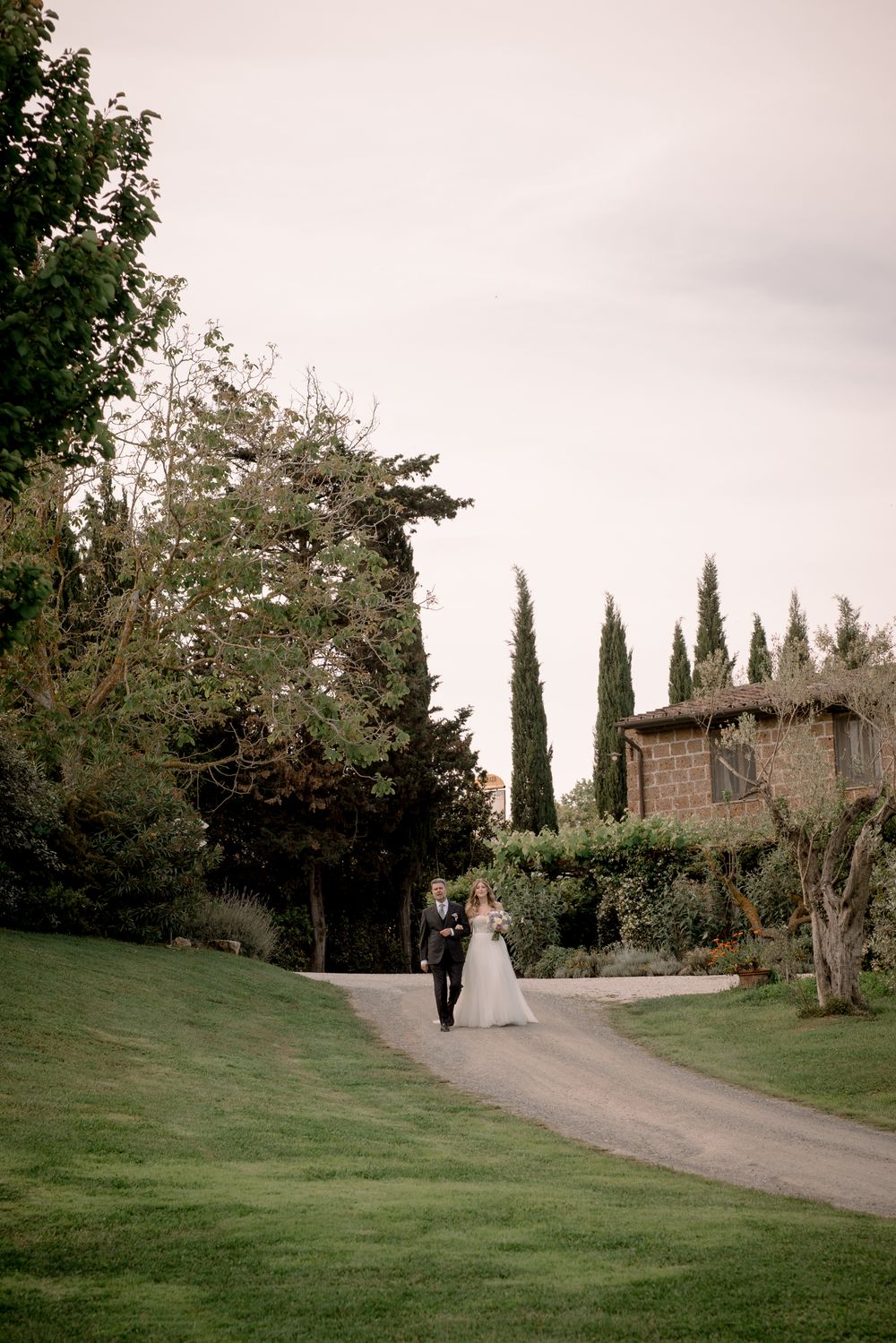 Bride and groom walking along a winding path at a Tuscan villa surrounded by cypress trees and lush gardens.