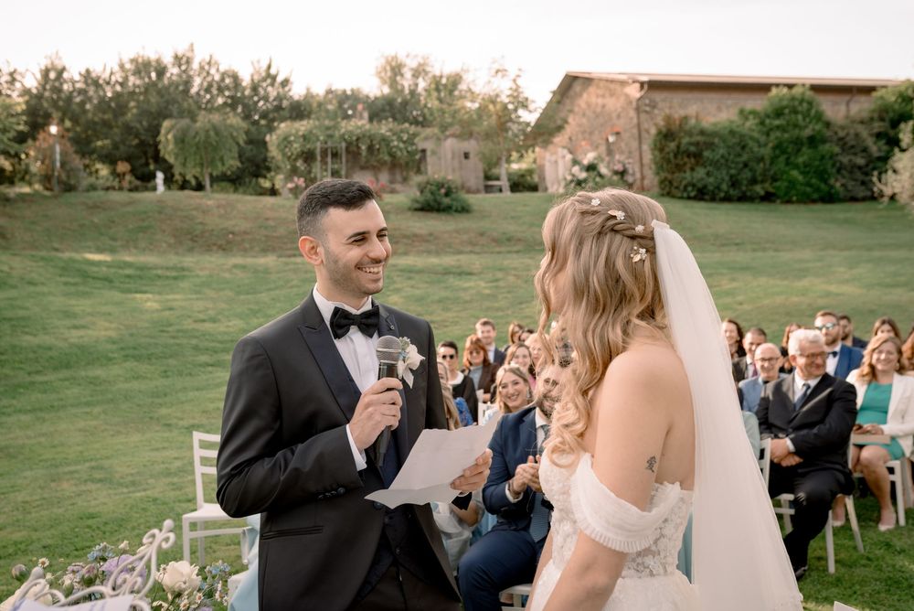 Groom smiling and reading vows to bride at an outdoor wedding ceremony with guests in background.