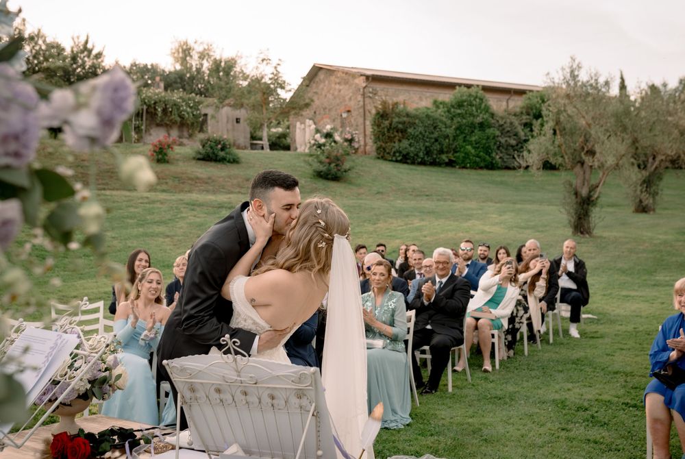Bride and groom share first kiss at outdoor wedding ceremony in Tuscany, guests applauding in background.