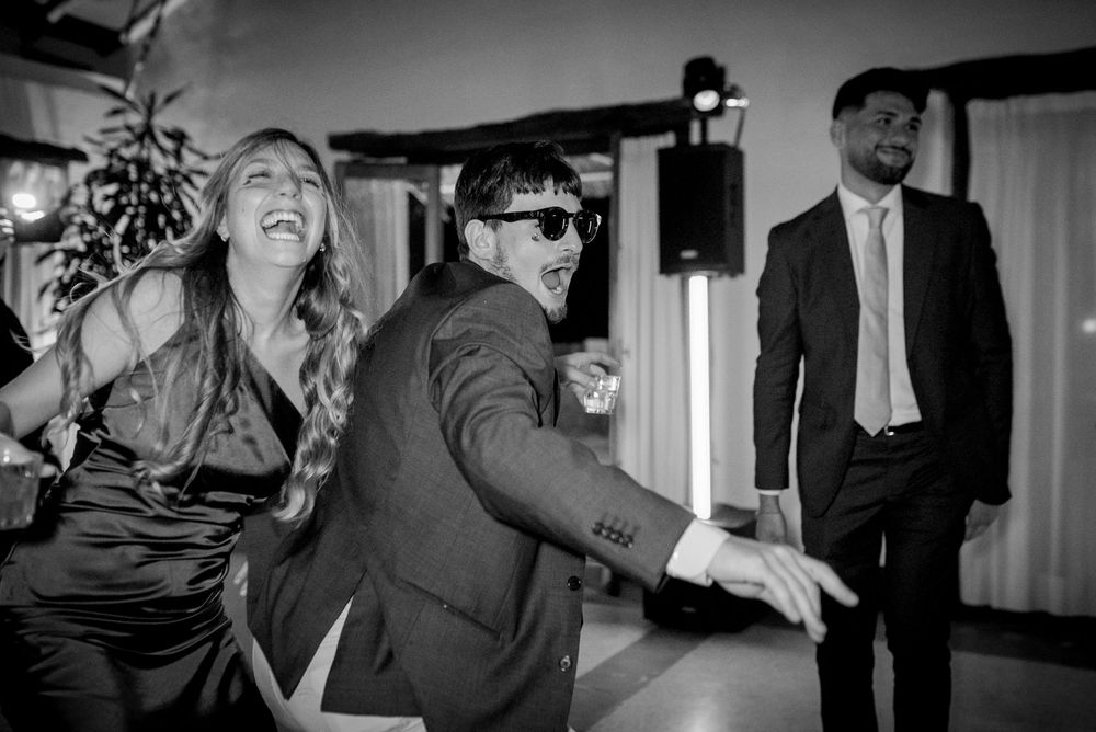 Black and white photo of guests dancing and laughing at a wedding reception in formal attire.
