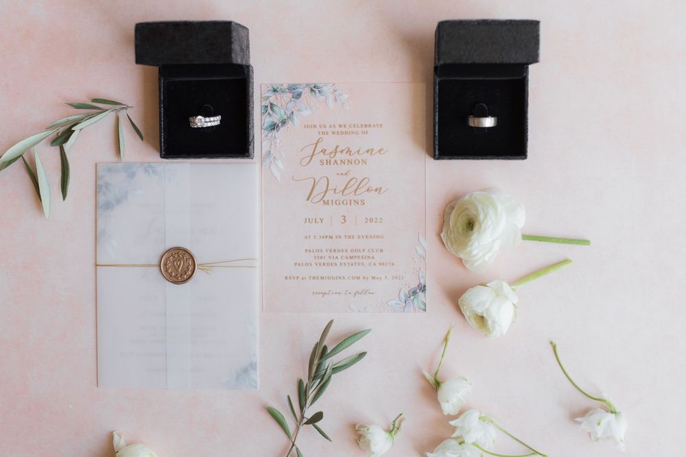 Elegant wedding flat lay with floral invitations, gold wax seal, white ranunculus flowers, and diamond rings in black boxes.