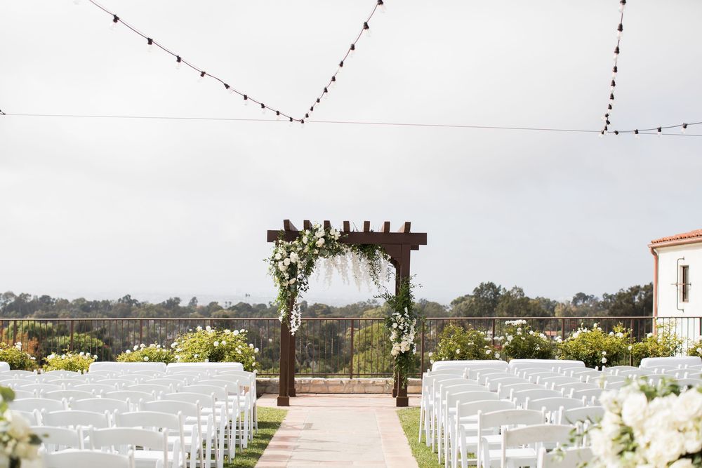 Elegant outdoor wedding ceremony setup with white chairs, floral arch, and string lights overhead.