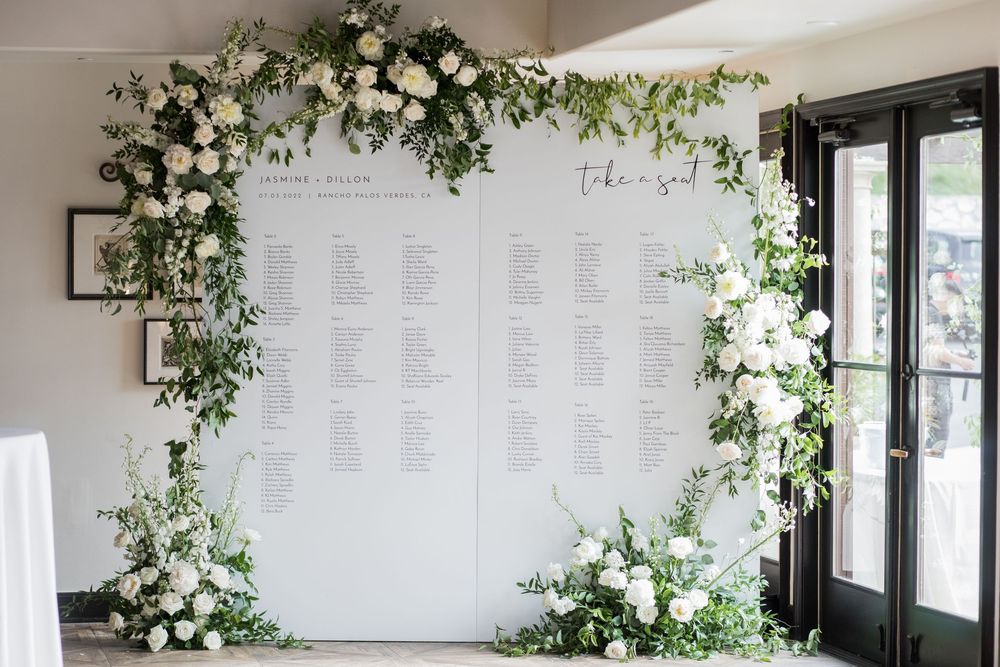 Elegant wedding seating chart display framed by lush greenery and white floral arrangements.
