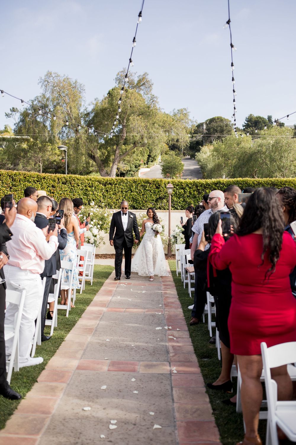 Bride and groom walk down outdoor wedding aisle surrounded by seated guests, string lights, and lush greenery backdrop.