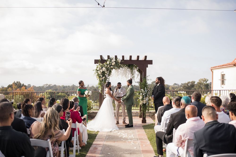 Outdoor wedding ceremony with bride and groom at floral arch altar, guests seated on white chairs in sunny garden venue.