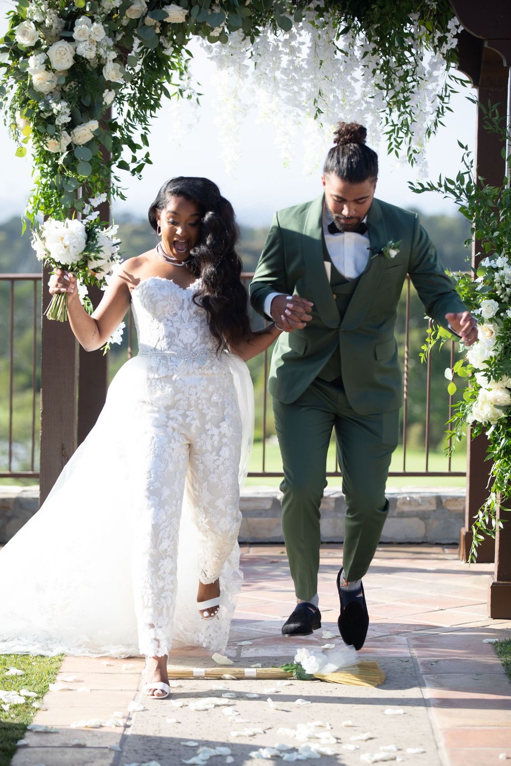 Joyful newlyweds exit ceremony under floral arch, bride in white lace jumpsuit, groom in green suit, breaking glass tradition.