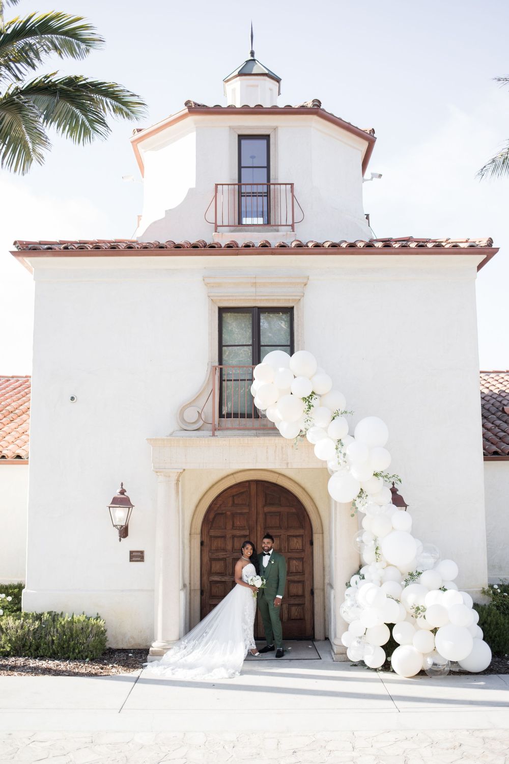 Bride and groom pose under white balloon arch at Spanish-style wedding venue entrance.