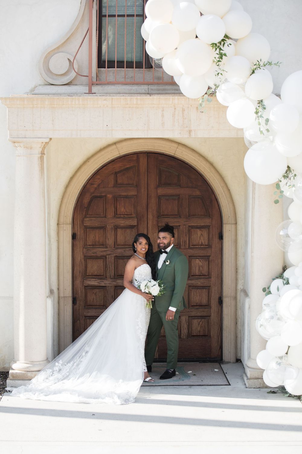 Bride and groom pose under white balloon arch at ornate wooden church doors on their wedding day.