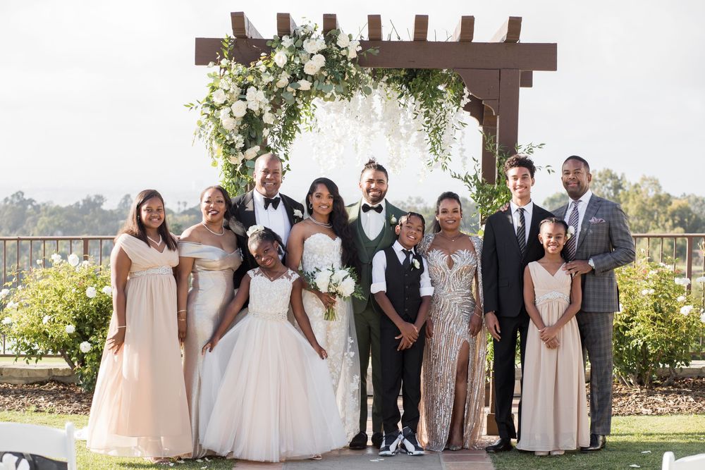 Wedding party poses under floral arch, bride in white gown with bridesmaids in blush dresses and groomsmen in suits.