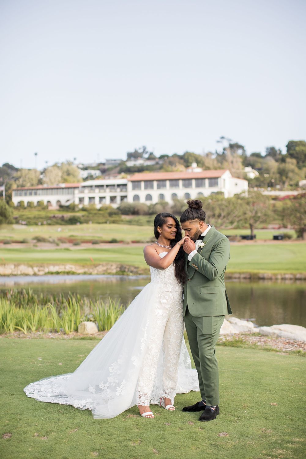 A groom in a green suit kisses his bride's hand as she wears a lace wedding gown, posing by a scenic pond.