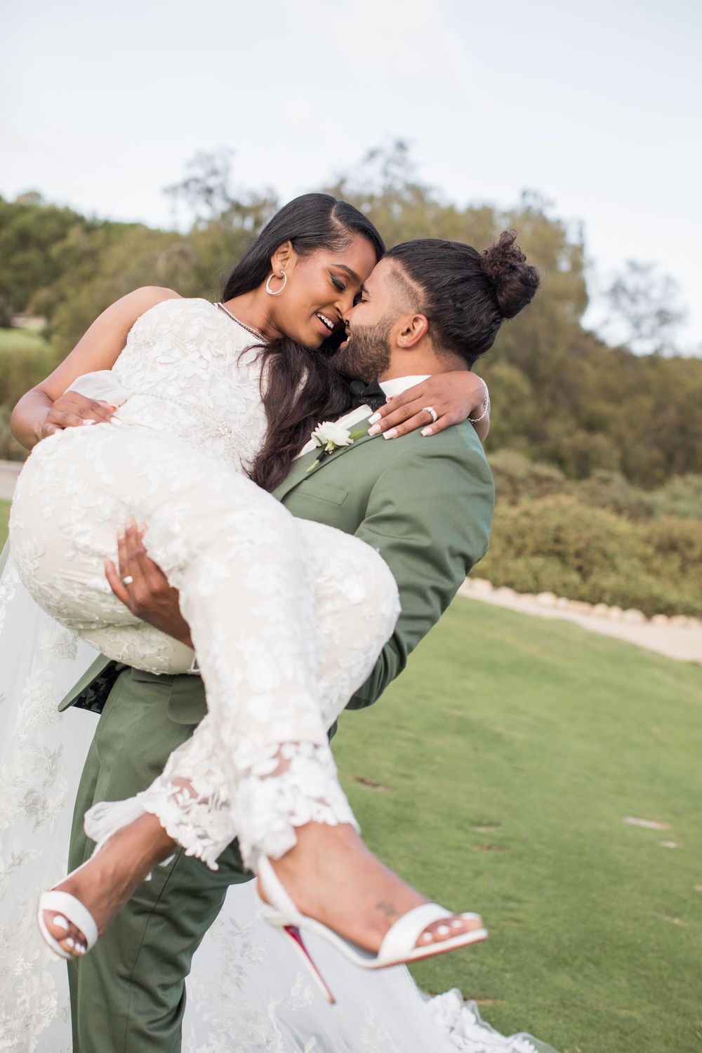 Groom in green suit lifts bride in white lace dress outdoors, sharing a romantic moment at their wedding.