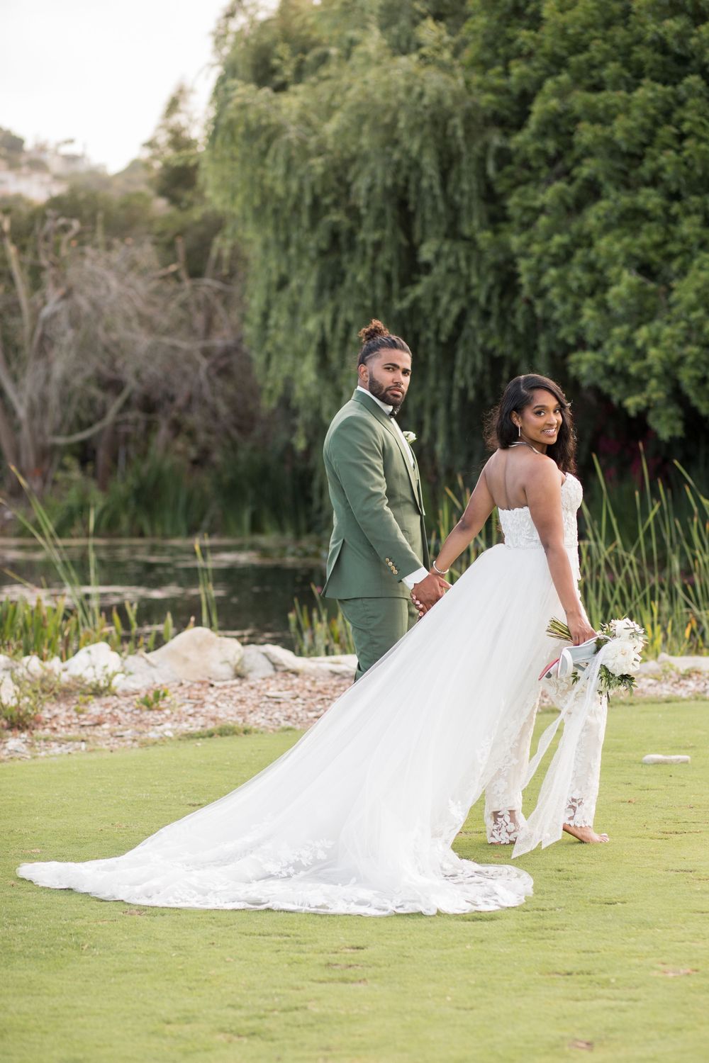 Bride in white gown and groom in green suit hold hands outdoors near a pond, posing for wedding portrait.