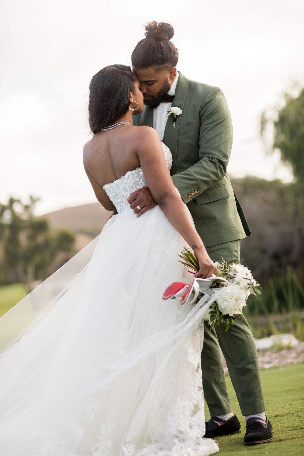 Groom in green suit kisses bride in strapless white ballgown outdoors, she holds white and pink bouquet.