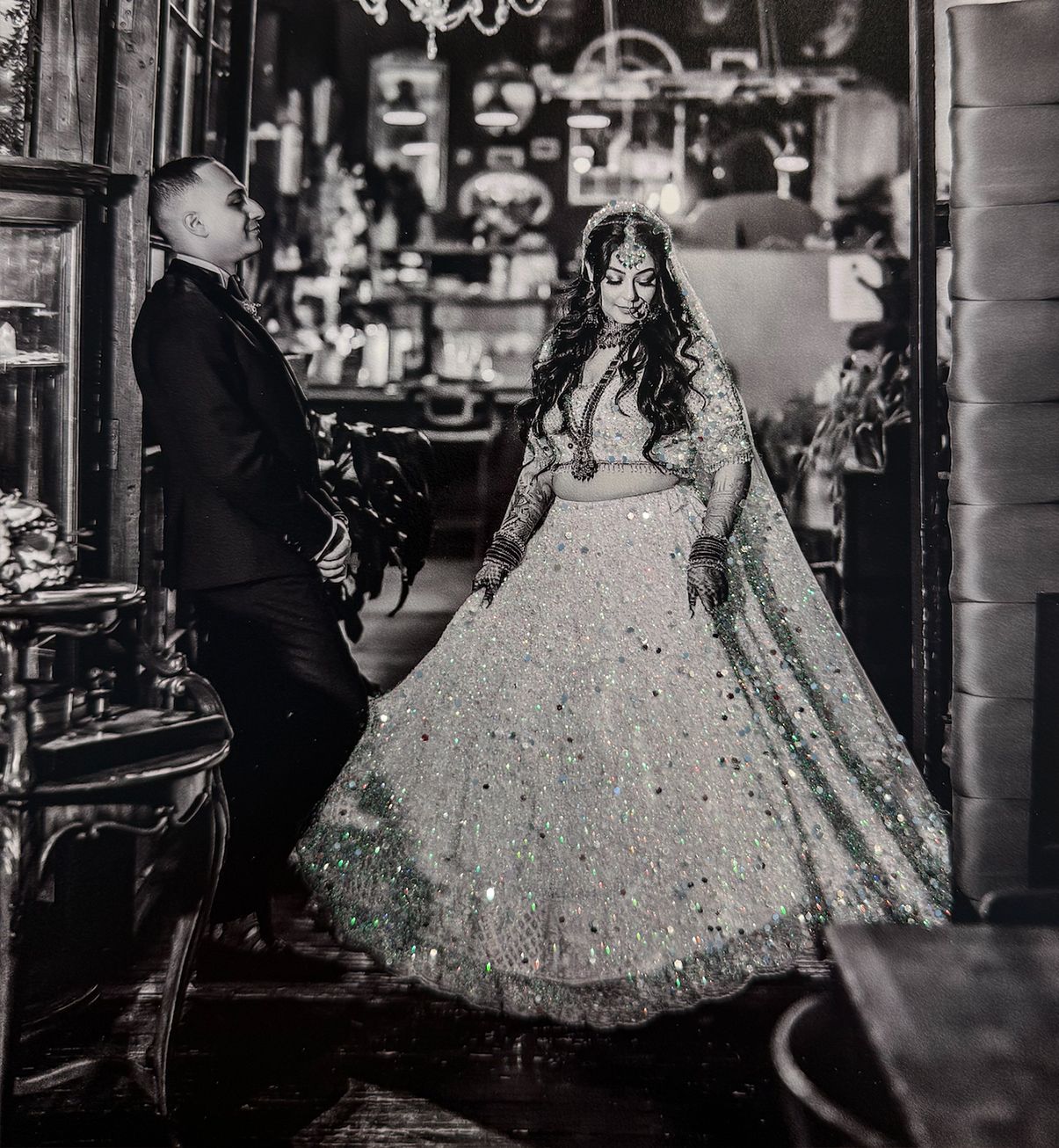 Black and white photo of a bride in a sparkling lehenga twirling before a groom in a suit at an elegant venue.