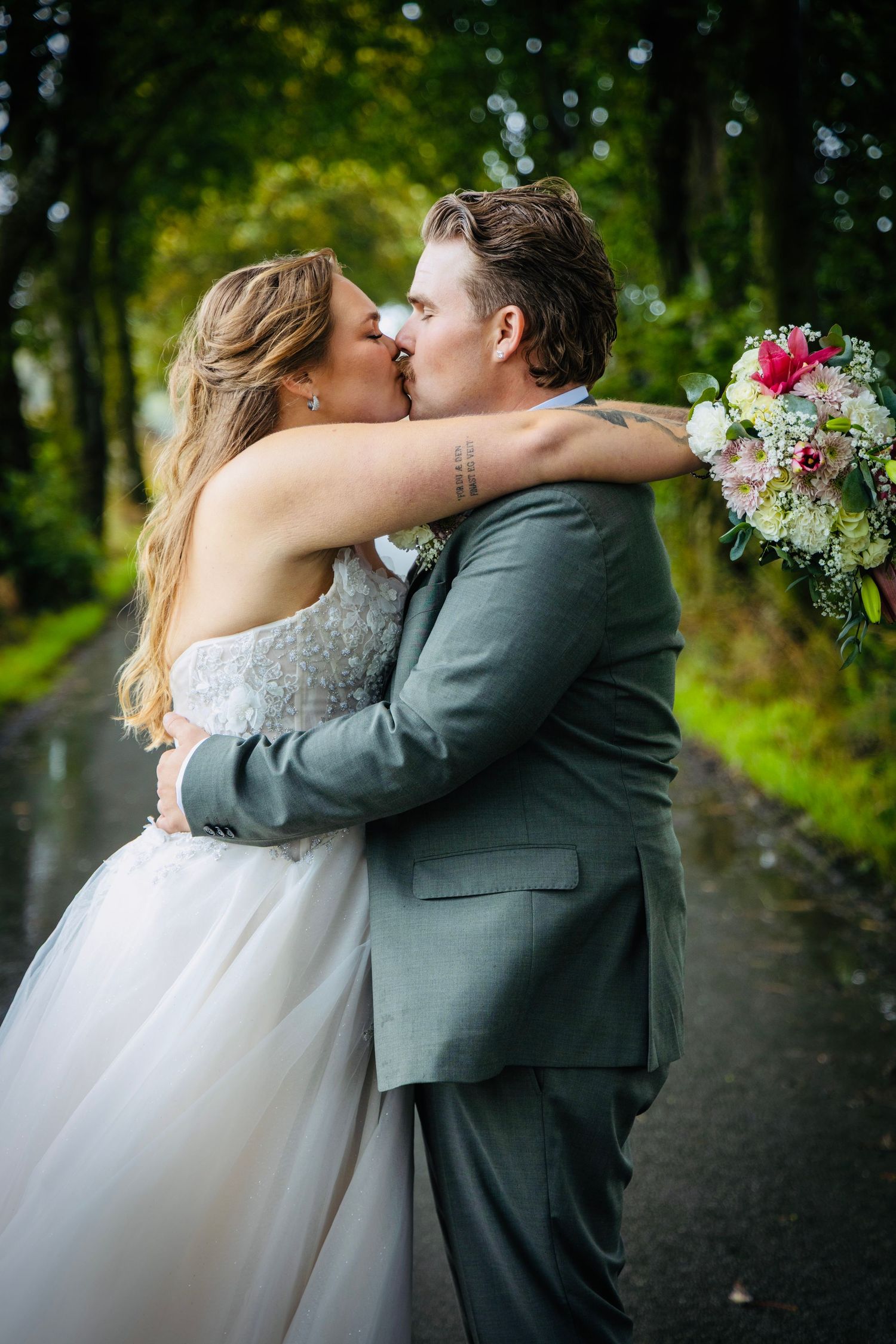 Bride and groom share a romantic kiss on a rain-soaked road, surrounded by lush green trees, holding a floral bouquet.