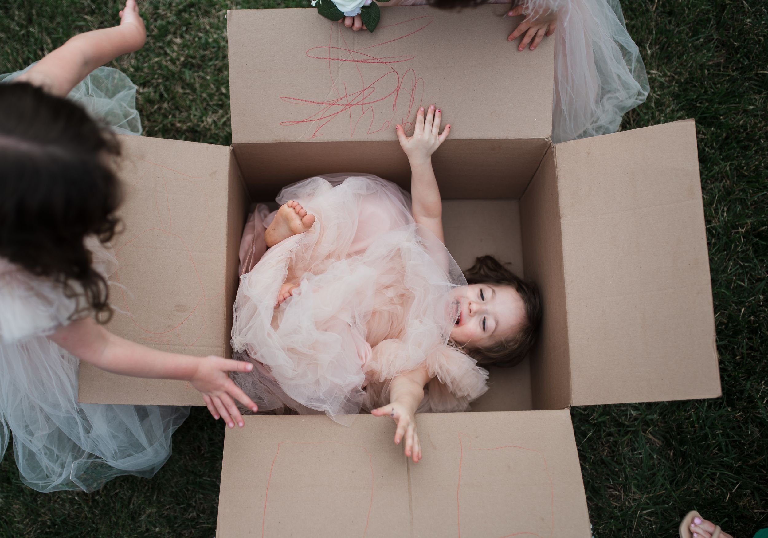Young girl in pink tulle dress sitting inside a cardboard box surrounded by other children in tulle skirts outdoors.