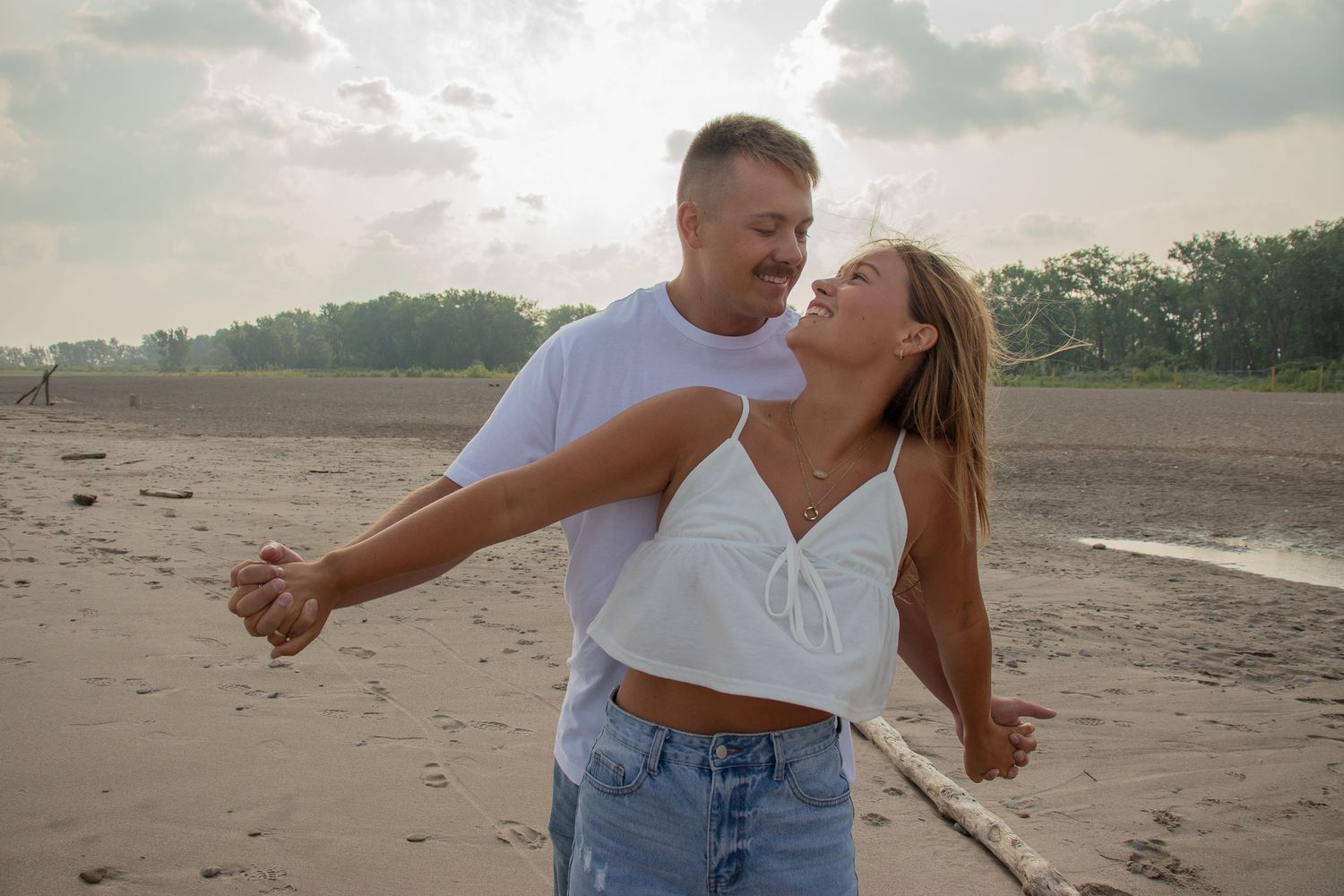 Couple dancing on a sandy beach at sunset, wearing white tops and jeans, smiling at each other.