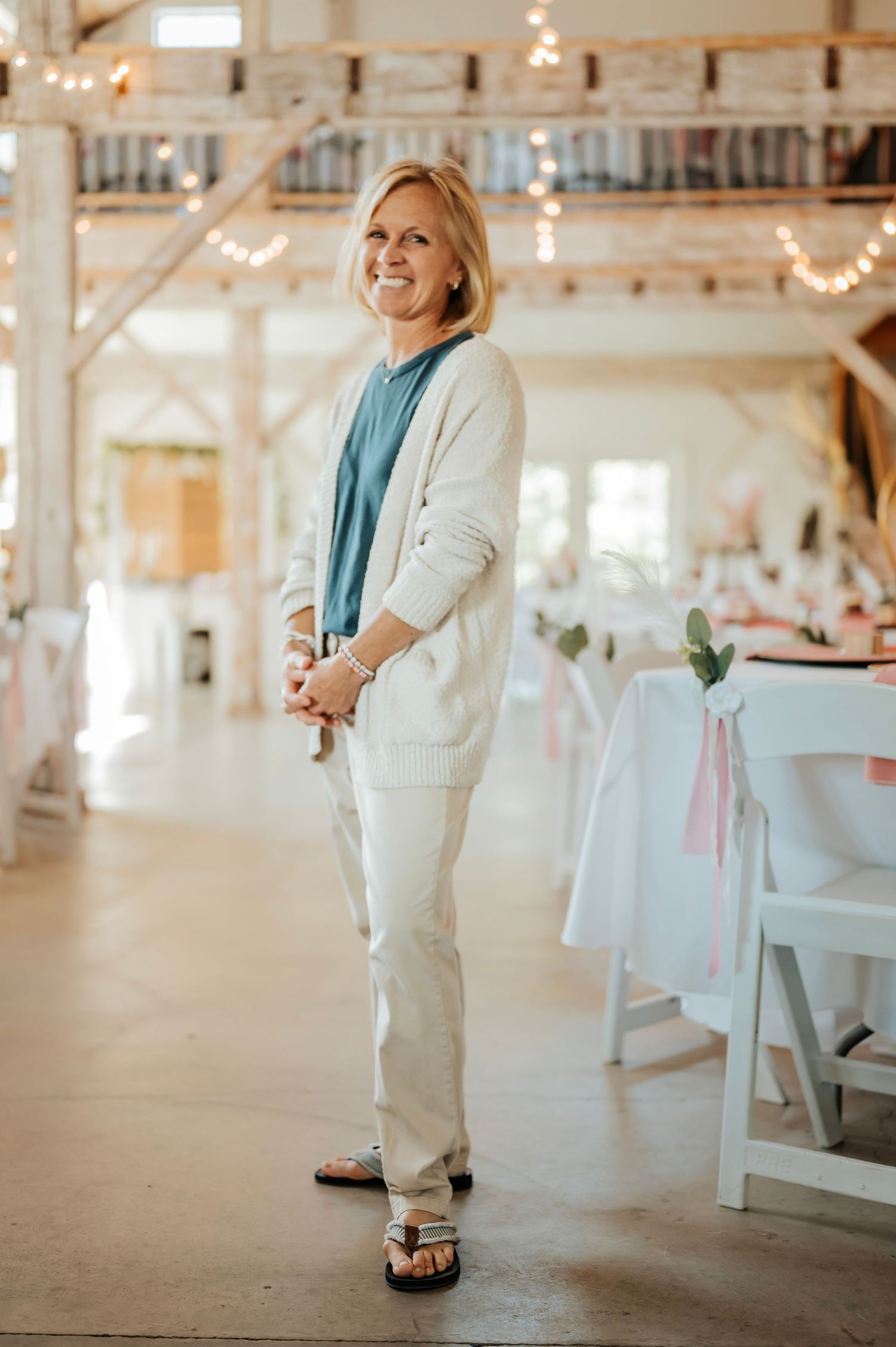 Smiling blonde woman in white cardigan and pants standing in elegant barn wedding venue with string lights.