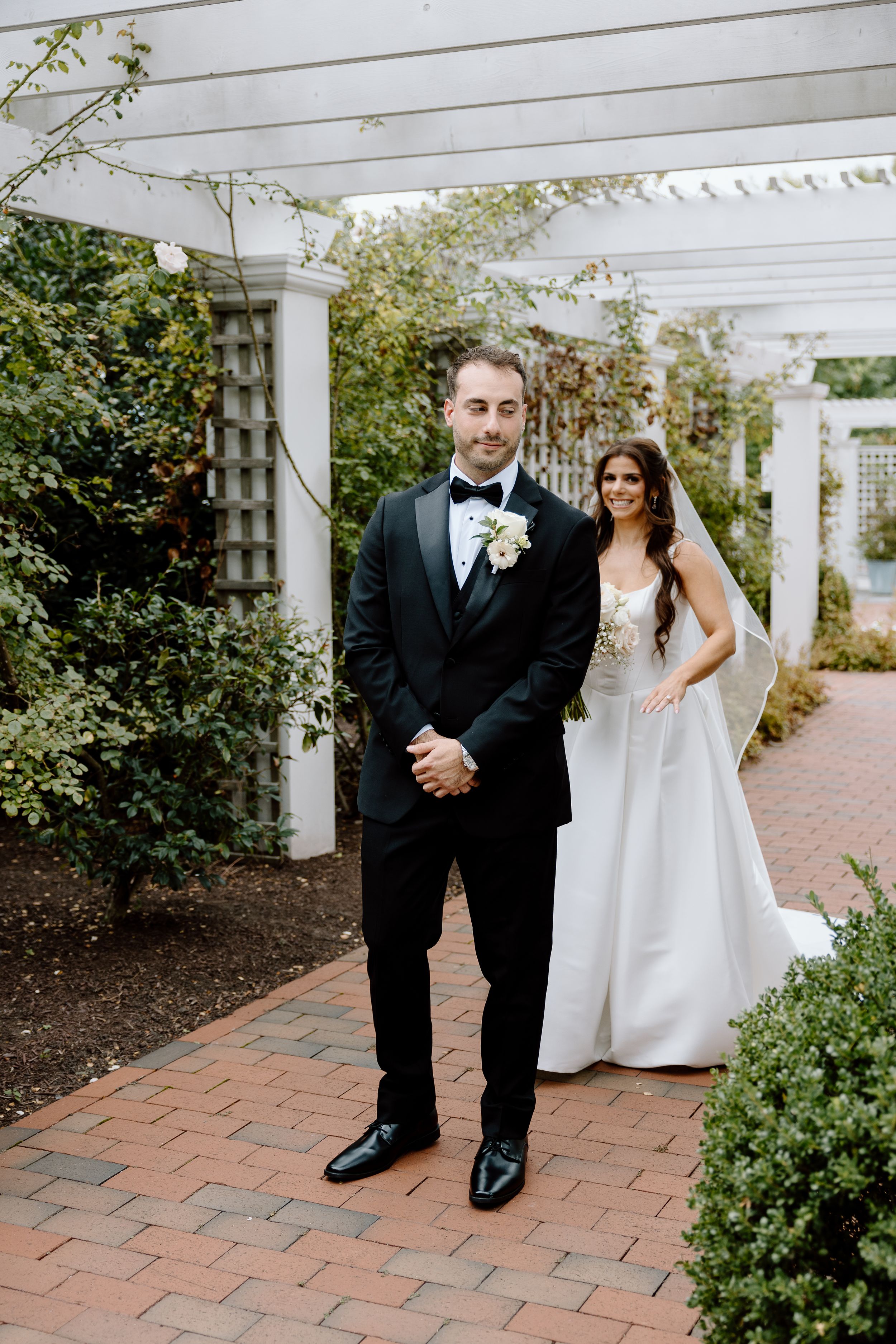 Bride in white gown and groom in black tuxedo pose under a white pergola surrounded by lush greenery on their wedding day.