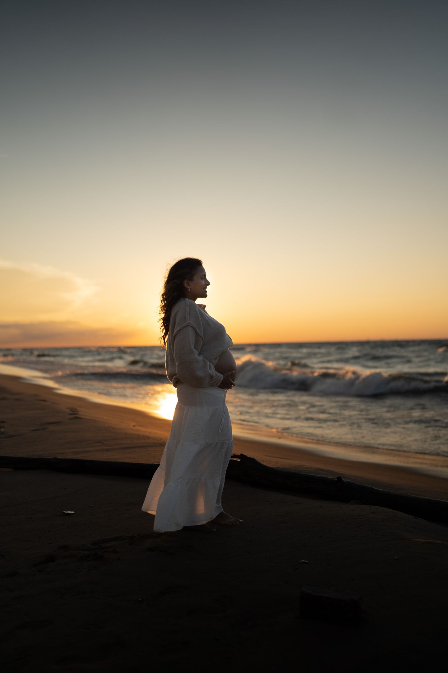 Pregnant woman in white dress standing on beach at sunset, cradling her belly while ocean waves crash behind her.