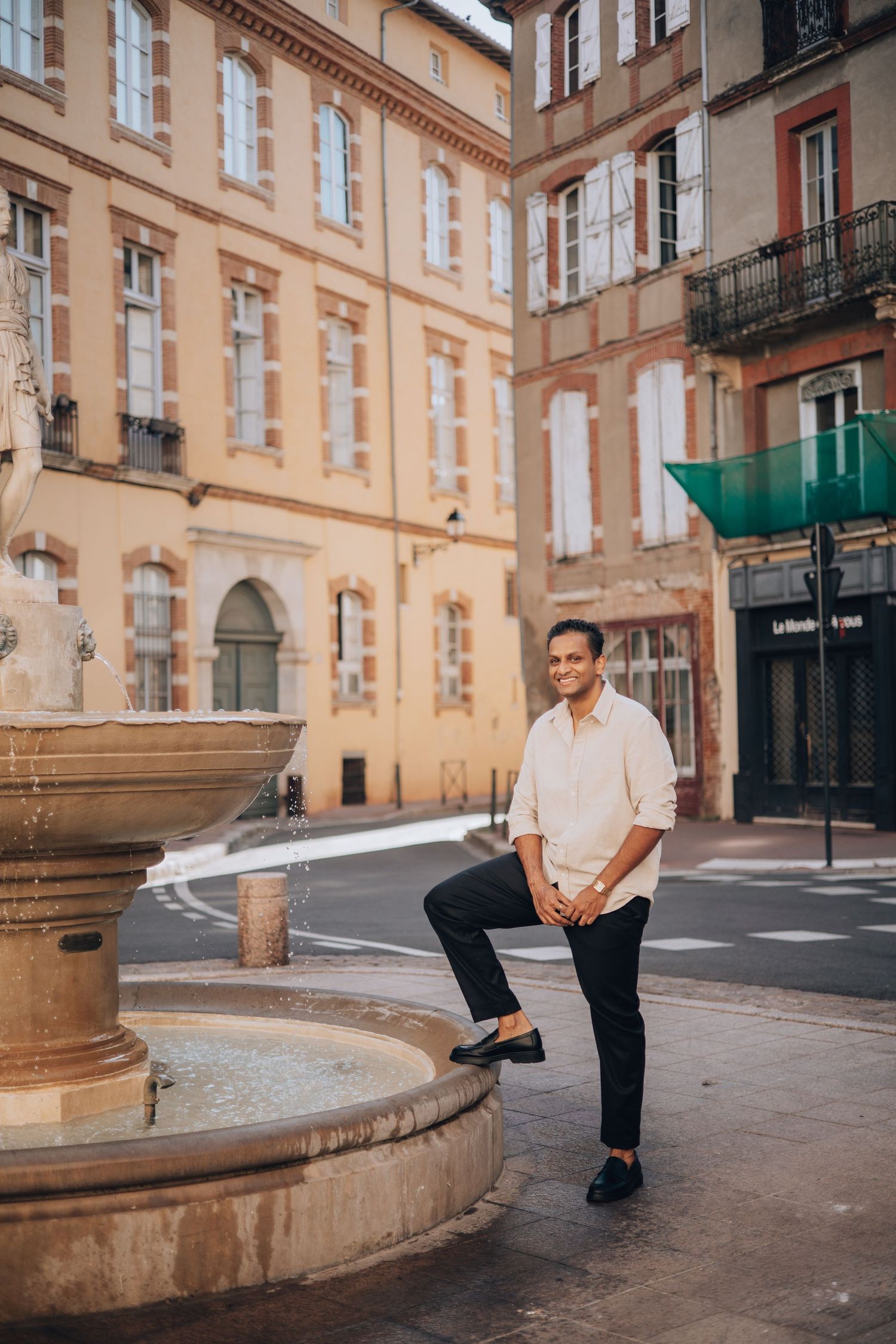 Man in white shirt posing by a stone fountain in a charming European city square with classic architecture.