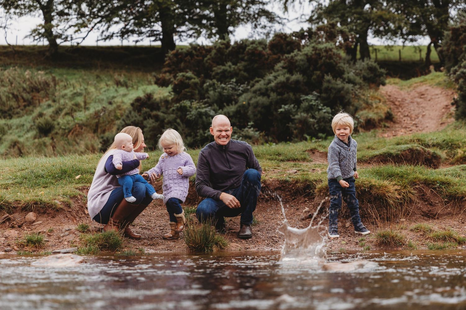 Family of four playing and splashing in a muddy puddle outdoors, smiling and enjoying nature together.