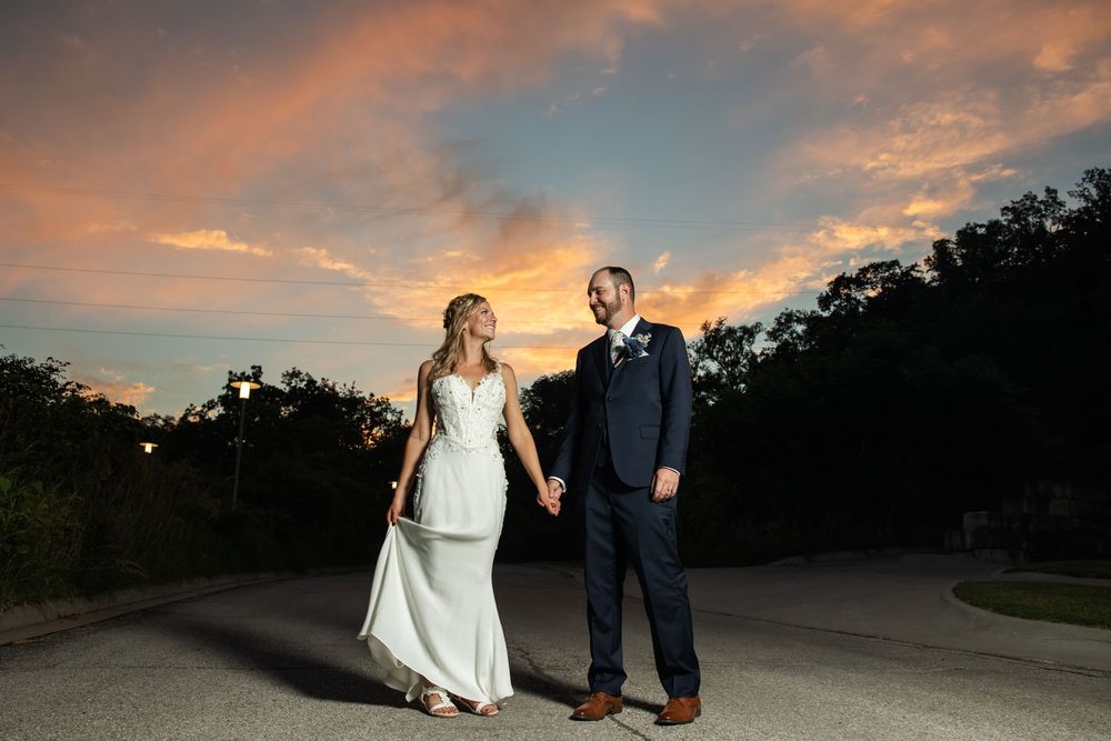 Bride and groom hold hands at sunset with vibrant orange clouds behind them on their wedding day.