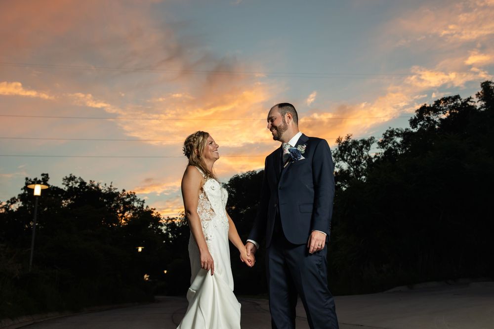 Bride and groom hold hands at sunset with vibrant orange and pink sky backdrop at outdoor wedding venue.