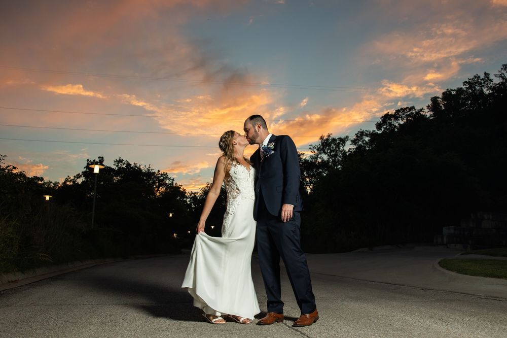 Bride and groom share a romantic kiss at sunset with a vibrant orange and blue sky backdrop on their wedding day.