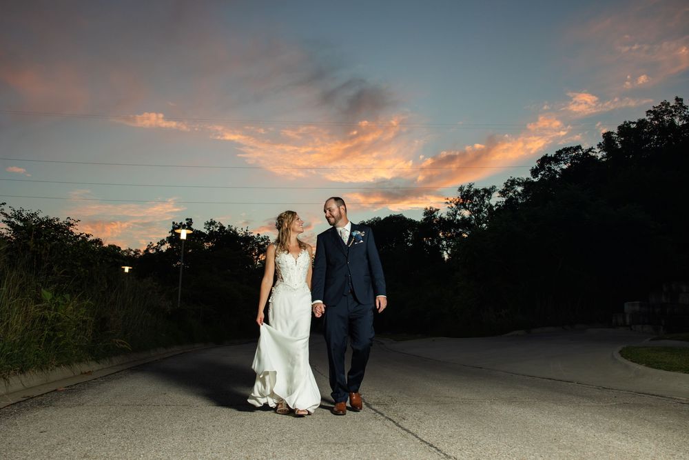 Bride and groom walking at sunset on a road, dramatic pink and orange sky behind them.
