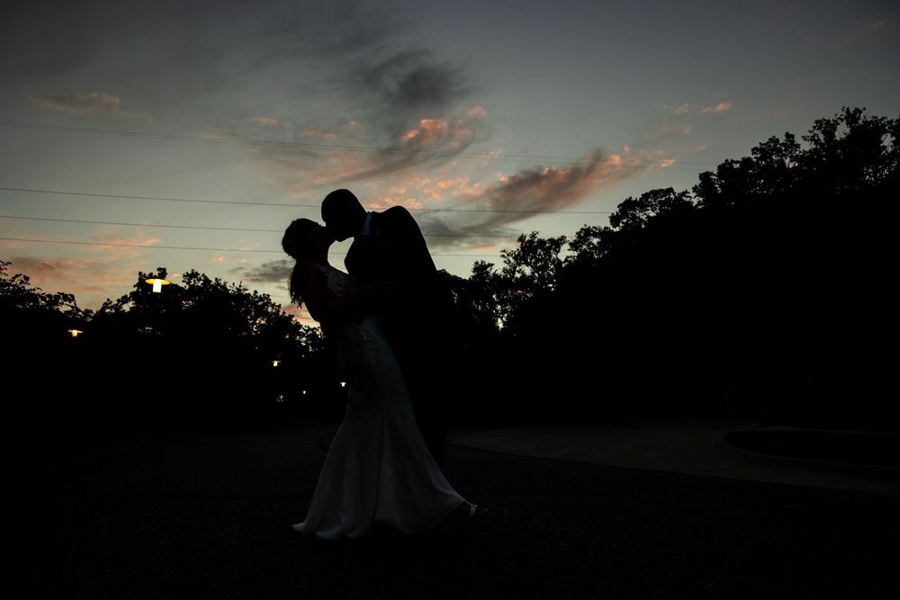 Silhouette of a couple sharing a romantic embrace at sunset, with a dramatic sky and dark tree line in the background.