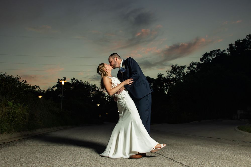 Bride and groom share a romantic dip kiss on a road at sunset, with a dramatic dusk sky backdrop.