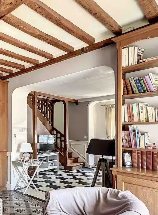Charming living room with exposed wood beam ceiling, checkered floor, built-in bookshelf, and elegant curved staircase.