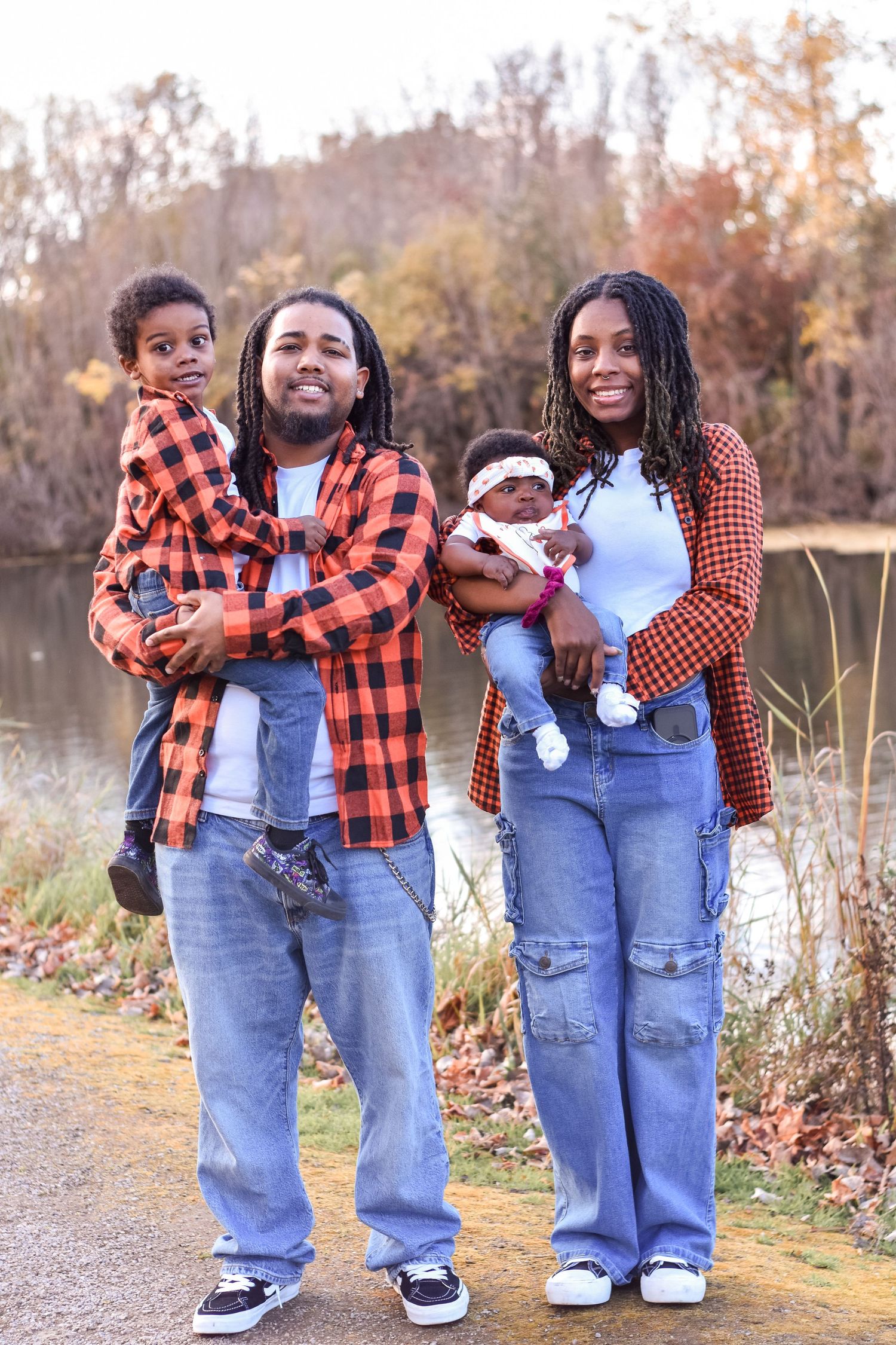 A joyful family of four poses outdoors in autumn, parents wearing matching plaid shirts and jeans, each holding a young child.