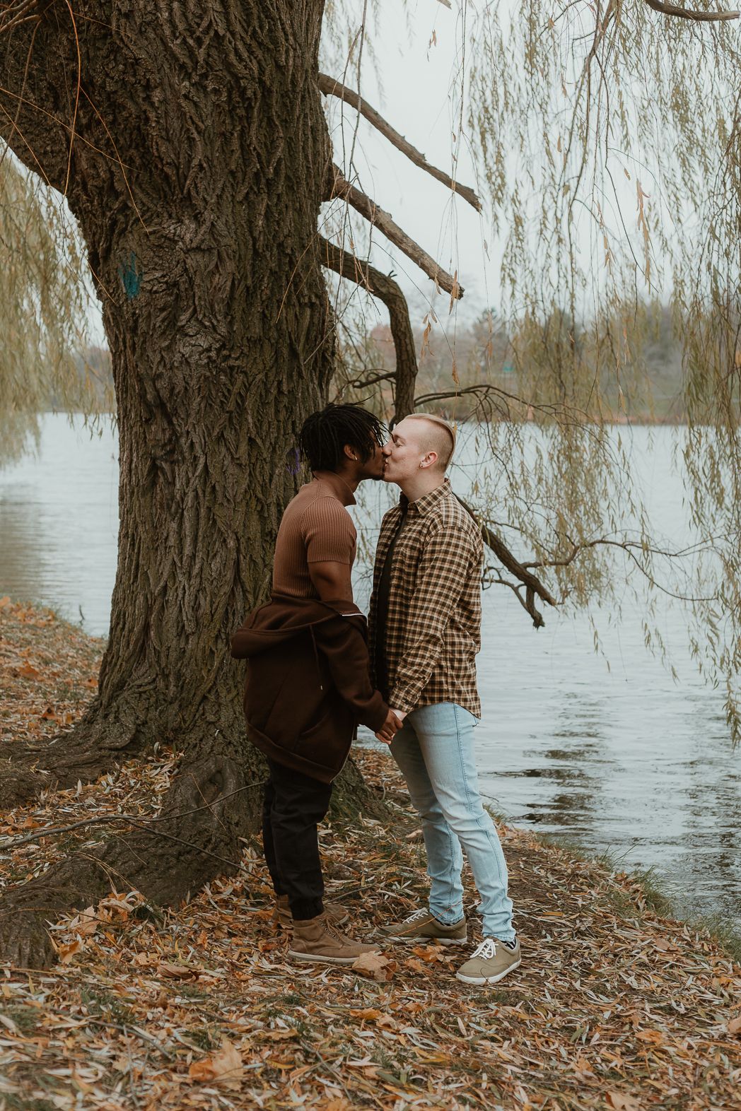 Interracial couple sharing a kiss under a willow tree by a lake in autumn.