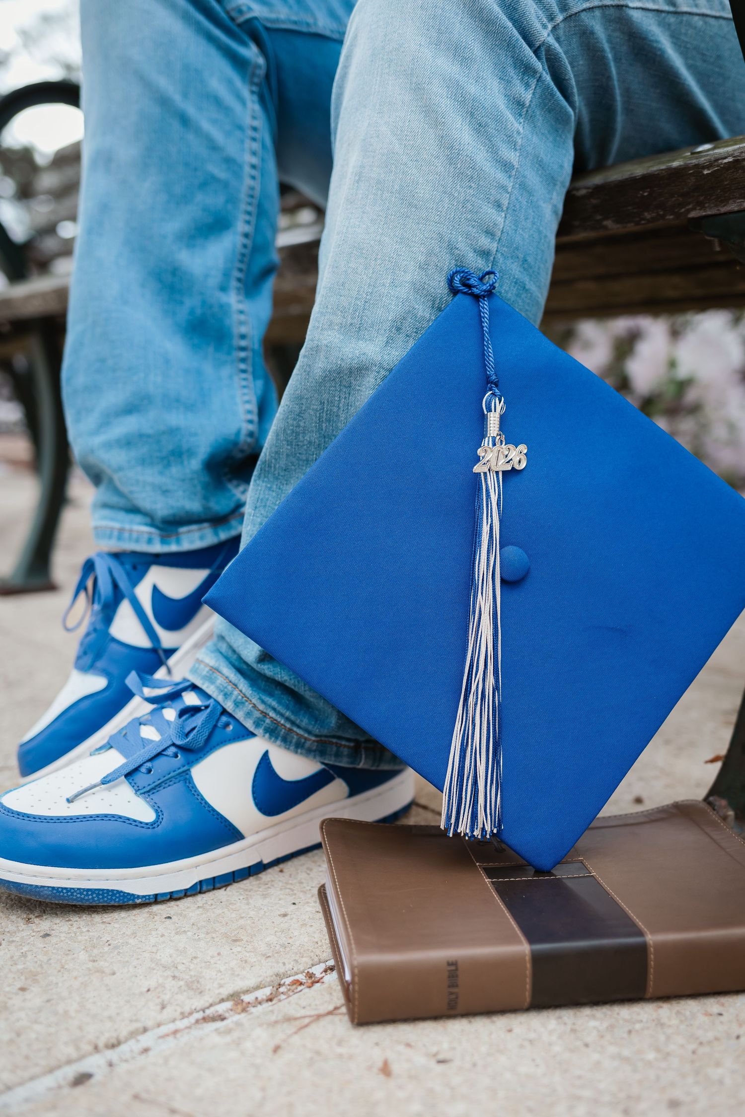 Blue graduation cap with 2026 tassel resting on a book beside blue and white Nike sneakers and jeans.