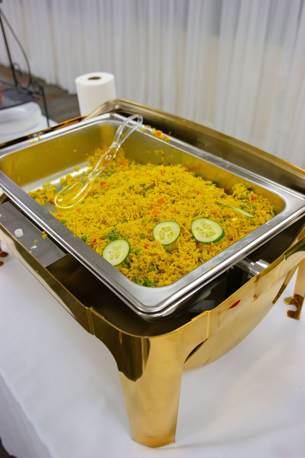 Golden chafing dish filled with yellow fried rice garnished with fresh cucumber slices at a buffet event.