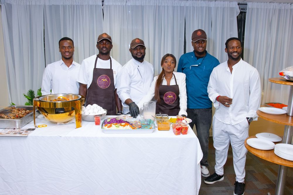Group of six people posing at a catering table with food and drinks at an indoor event.