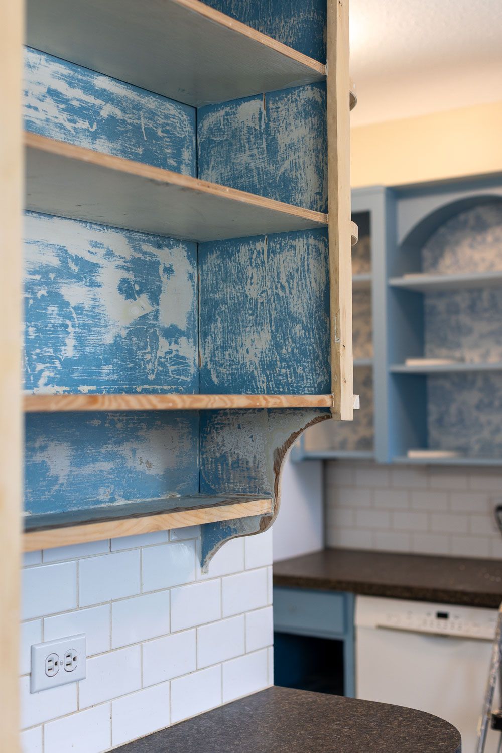 Rustic kitchen cabinet with distressed blue paint, wooden shelves, white subway tile backsplash, and dark countertop.
