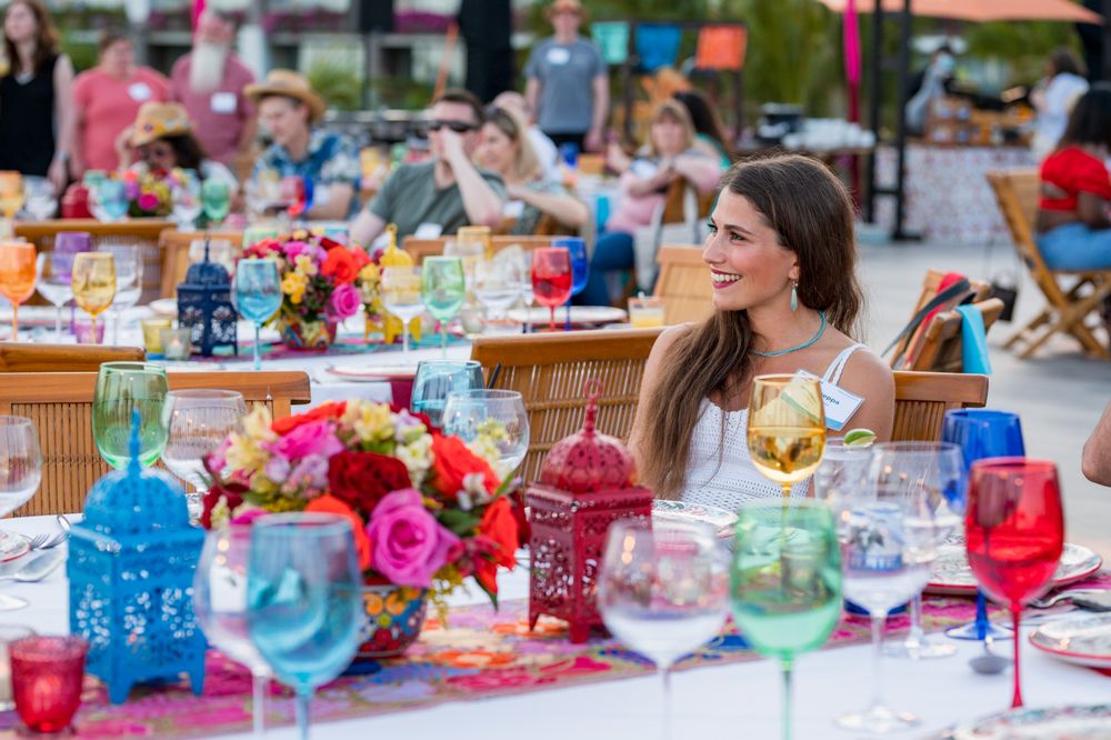 Woman smiling at colorful outdoor dinner party with vibrant glassware, floral centerpieces, and festive table decorations.