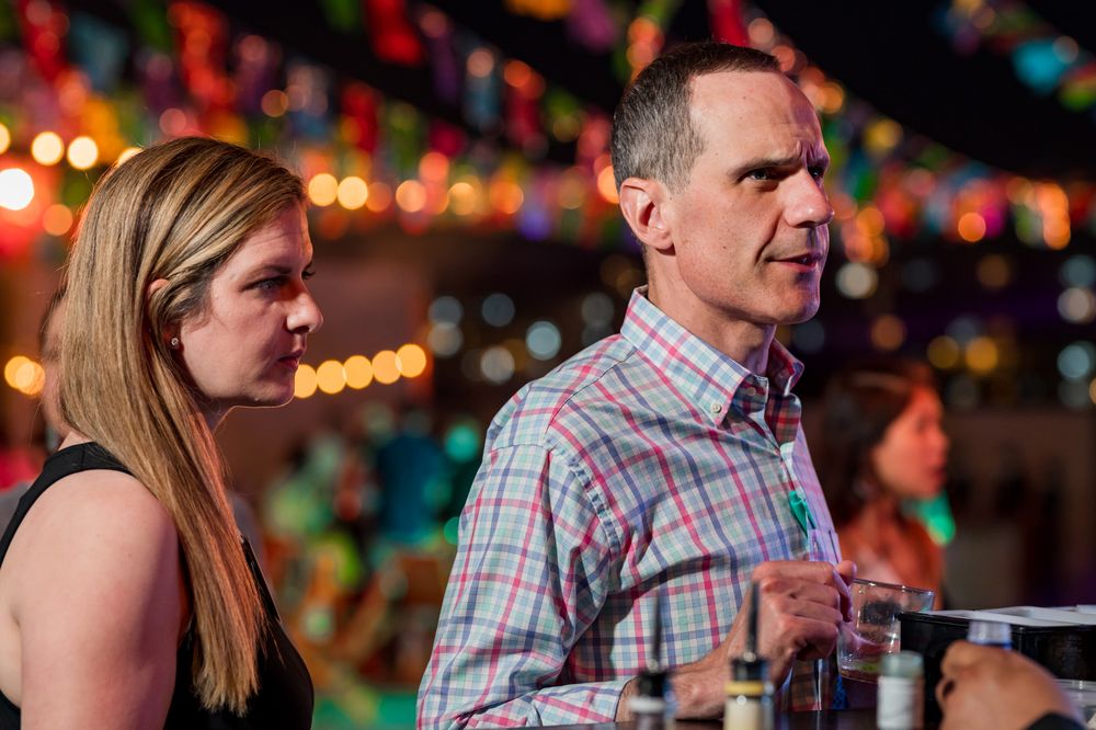 A man and woman stand together at a colorful, festively lit indoor event with string lights in the background.