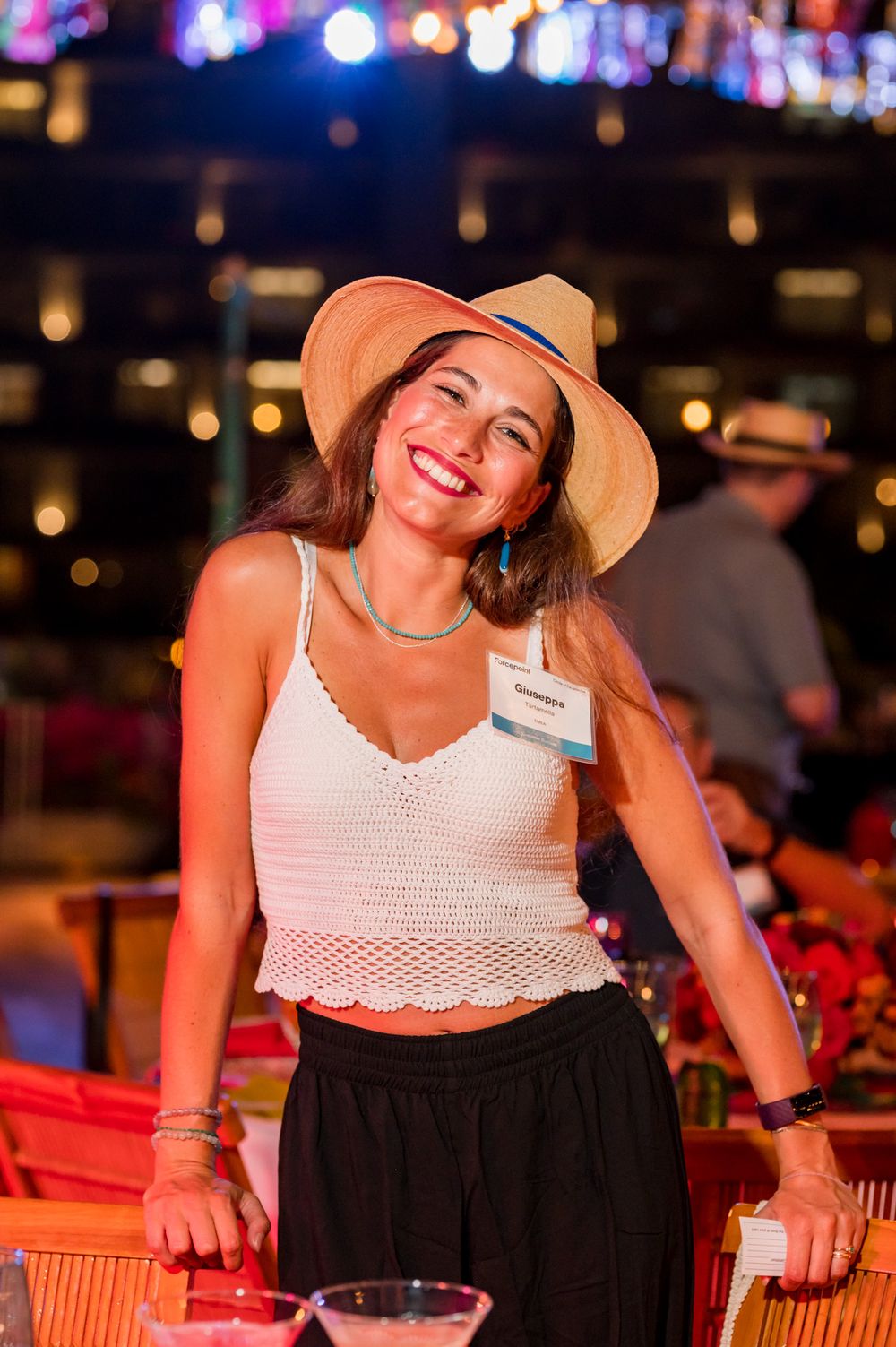Smiling woman in straw hat and white crochet top at a colorful outdoor evening event.