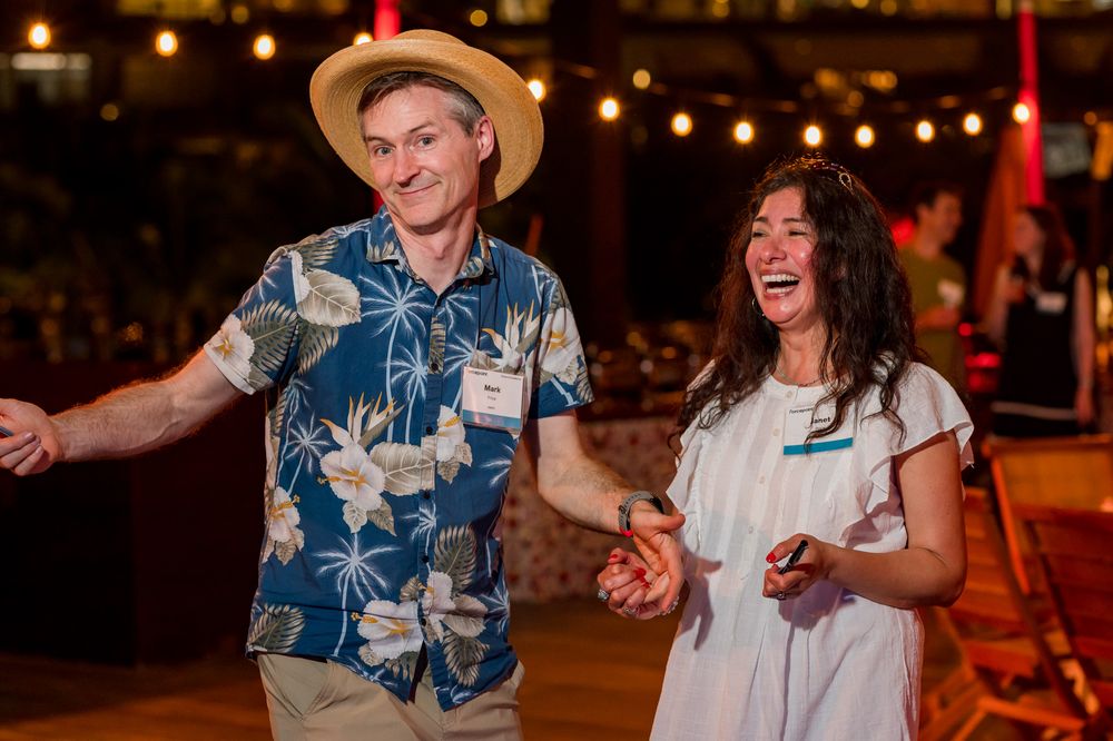 A man in a Hawaiian shirt and straw hat dances with a laughing woman in white at an evening event.