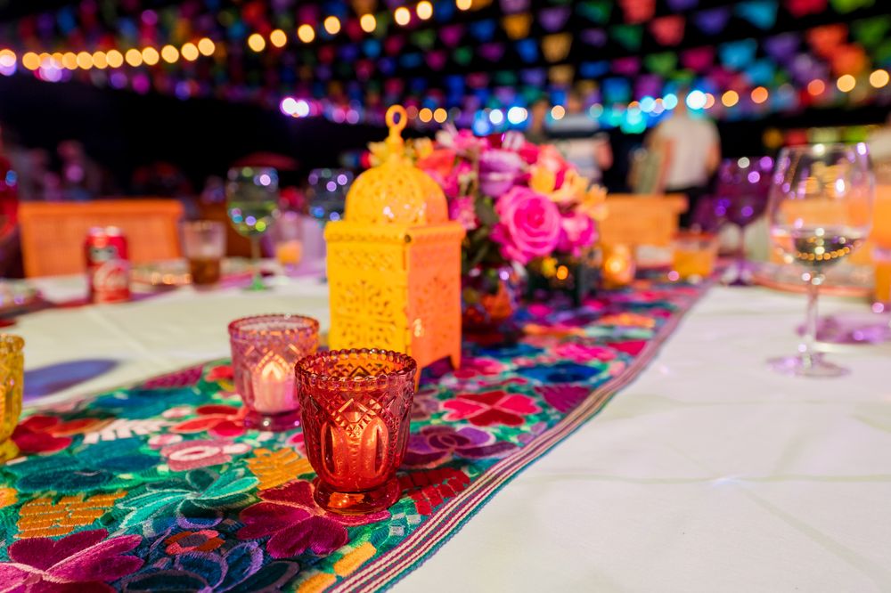 Colorful Mexican fiesta table setting with floral runner, gold lantern centerpiece, and vibrant candle holders under string lights.
